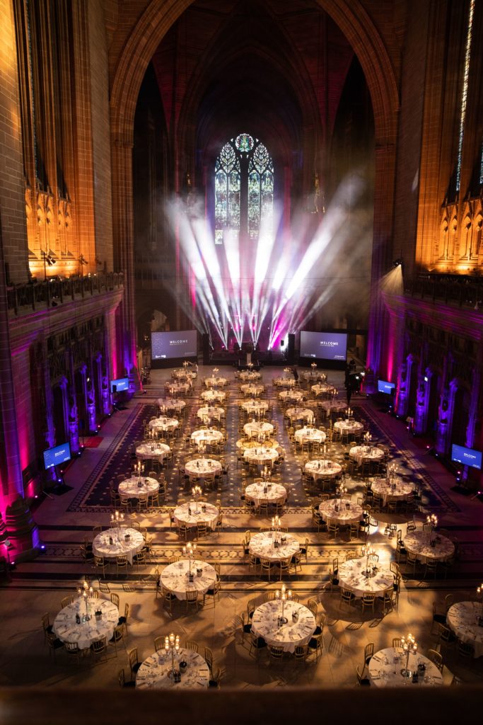 A set up banquet in a spacious cathedral . Lit up with white lights and purples and pinks.